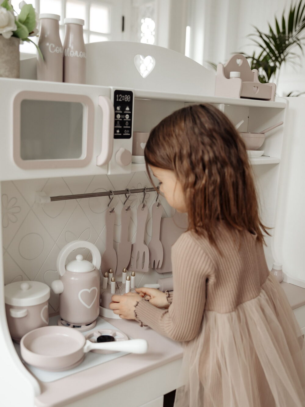 Девочка в бежевом платье и милой улыбкой ставит игрушечный тортик на фоне бело-бежевой детской игровой кухни.