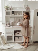 Девочка в бежевом платье и милой улыбкой держит в руках игрушечный тортик на фоне бело-бежевой игровой кухни.
