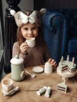 Девочка в милой маске зайчика и коричневой кофте играет с детским игровым набором для чаепития.