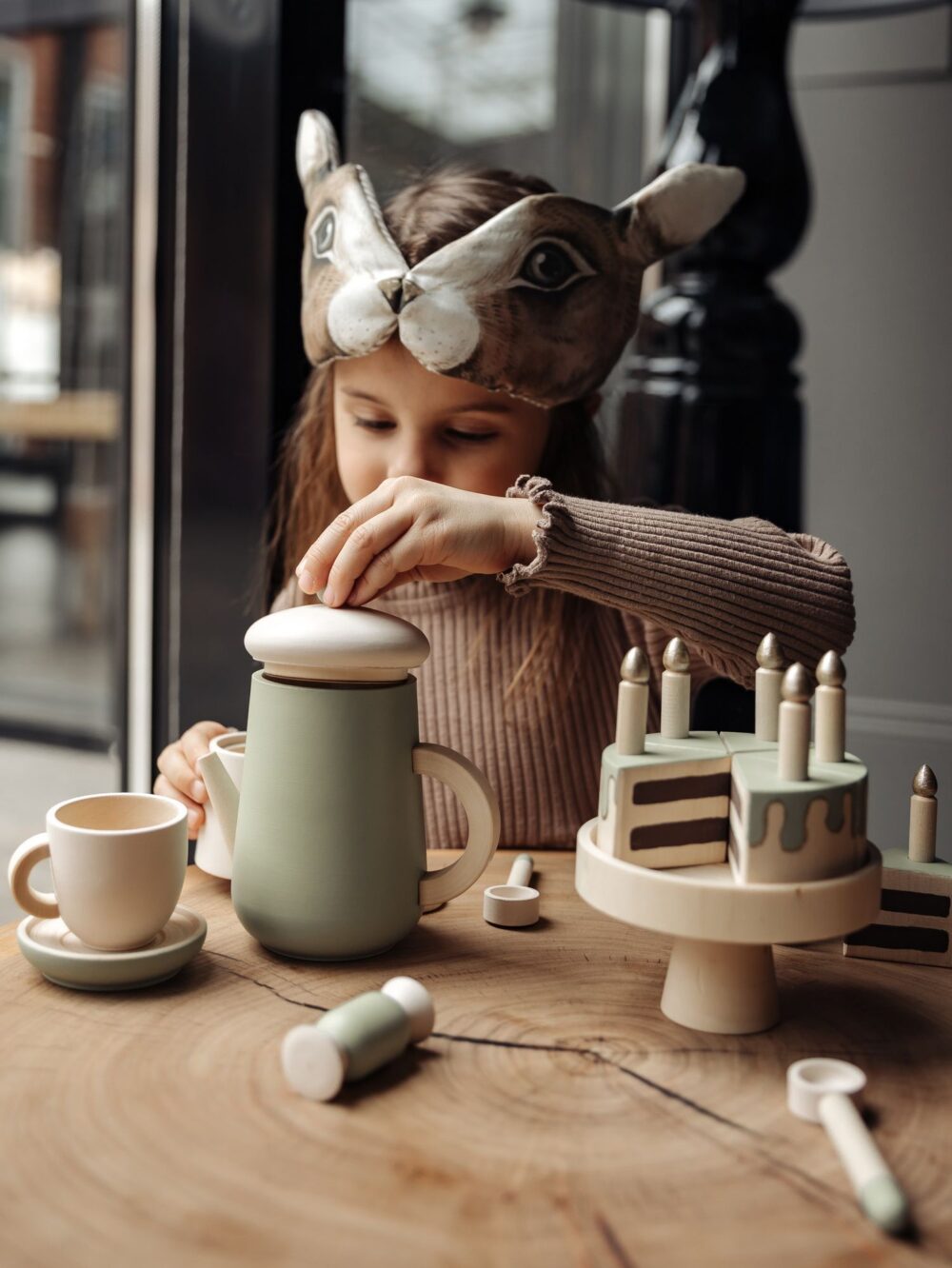 Девочка в милой маске зайчика и коричневой кофте играет с детским игровым набором для чаепития.