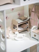 На нежном розовом фоне, красивый кукольной домик бело-бежевого оттенка, внутри домика горит мягкий свет. На обоях домика розовые бабочки и сиреневые цветы. Хорошо видны детали кукольный мебели. Близкий вид отдельной комнаты