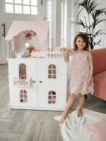 Красивый кукольный домик с дверями, Девочка облокотившись позирует на фоне. На домике нарисованы милые зайки. Вид с закрытыми дверками, внутри лежит кукла барби