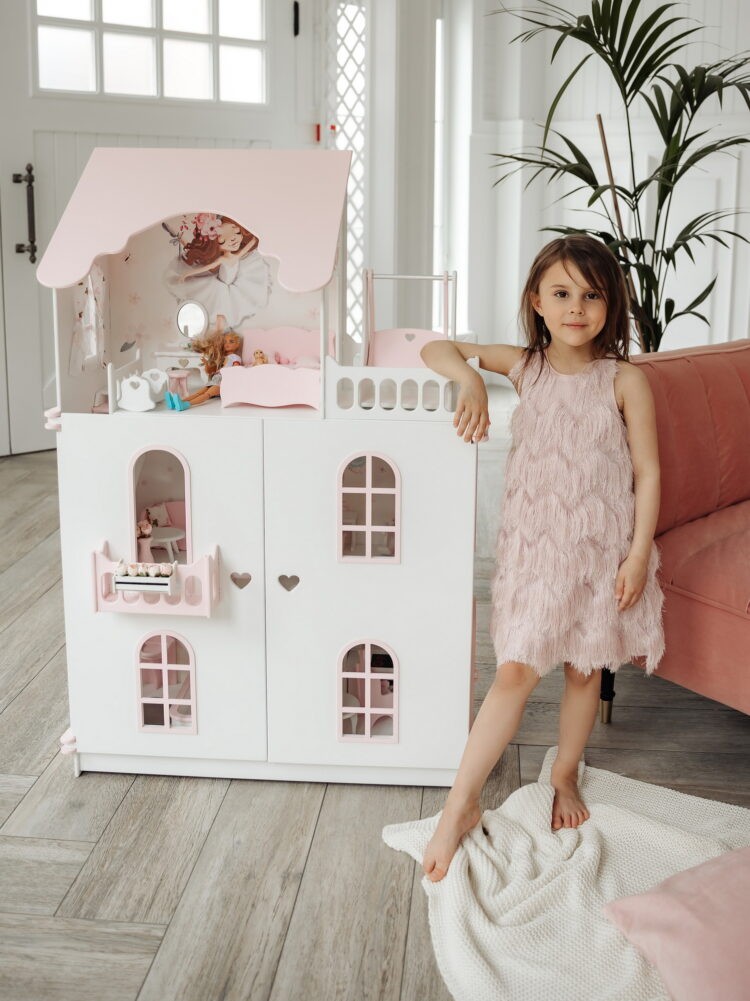 Красивый кукольный домик с дверями, Девочка облокотившись позирует на фоне. На домике нарисованы милые зайки. Вид с закрытыми дверками, внутри лежит кукла барби