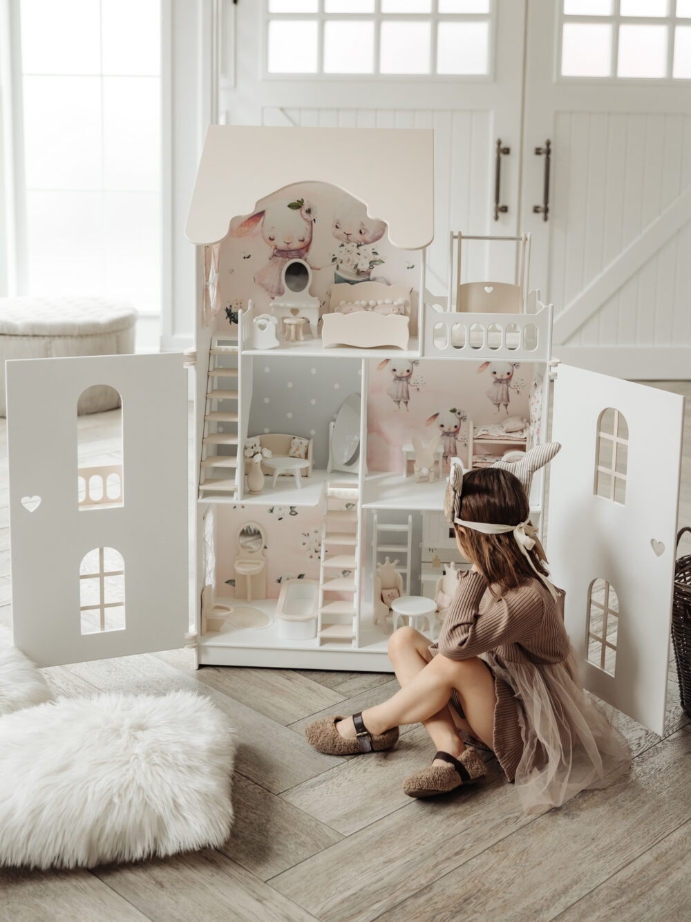 Красивый кукольный домик с дверями. Девочка сидит на полу и играет. На домике нарисованы милые зайки. Вид с открытыми дверками, внутри лежит кукла барби.