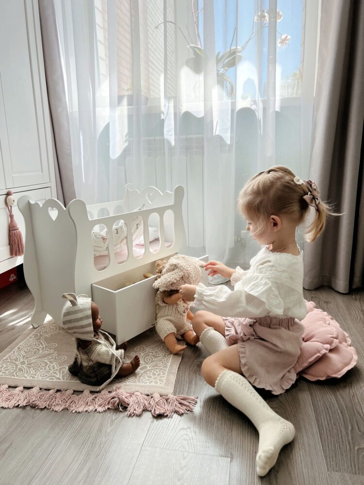 Белая люлечка для кукол в красивой детской комнате на полу. Рядом с ней сидит девочка, выдвинув ящик и играет с куклами.
