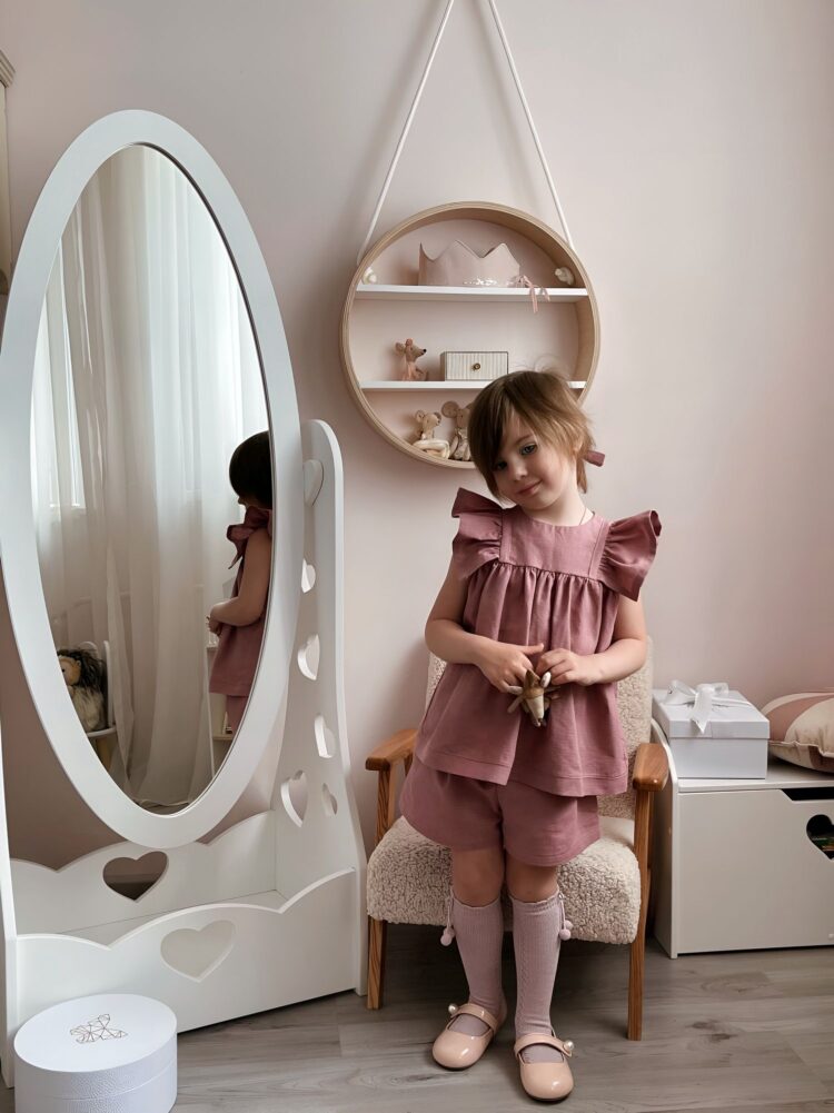 Красивая комната тепло-розового цвета. На фоне стоит девочка в красивом жемчужном платье рядом с белым детским напольным зеркалом.
