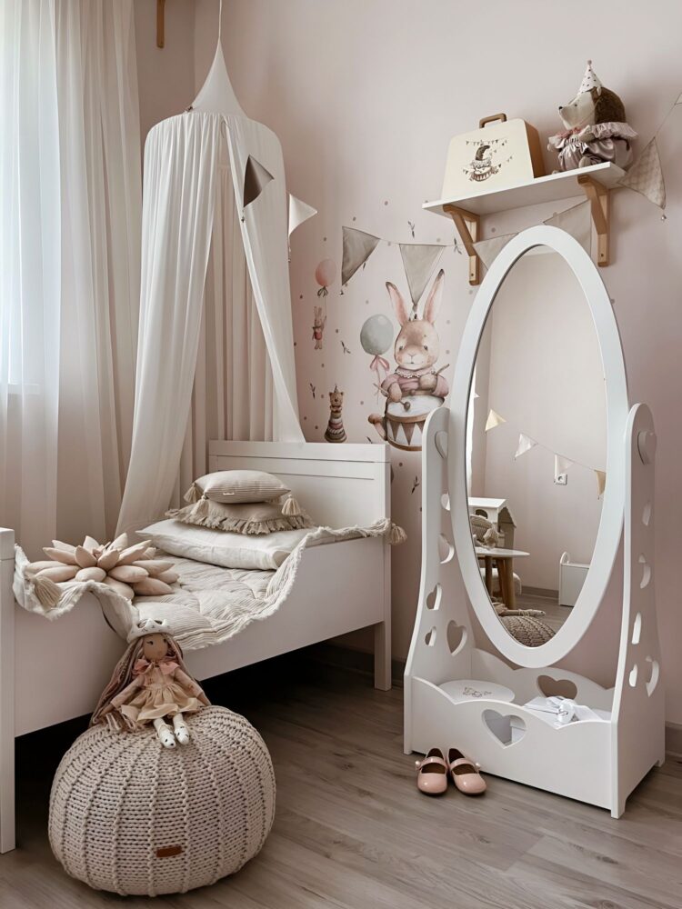 Красивая комната тепло-розового цвета с забавными зайками на стене. Рядом с детской кроватью стоит белое детское напольное игровое зеркало и вязанный пуфик с красивой куклой на нем.