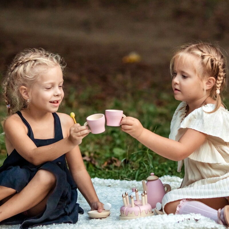 Две девочки в платьях на природе играют в чаепитие при помощи детского игрового набора для чаепития.
