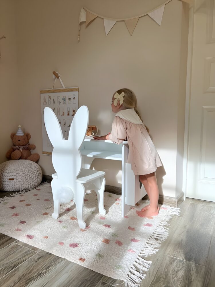 Милая девочка в розовом платье стоит облокотившись на детский белый столик со стулом в форме зайчика. Красивая комната с бежевыми обоями. В углу плюшевый мишка и игрушечный домик