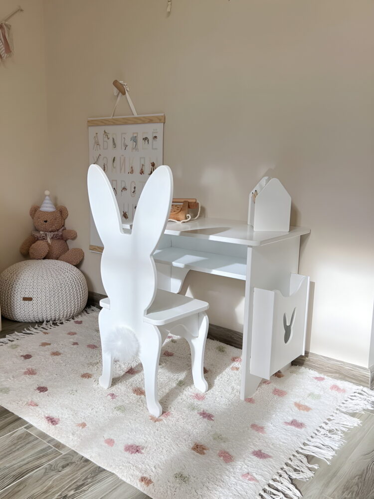 Детский белый столик со стулом в форме зайчика. Красивая комната с бежевыми обоями. В углу плюшевый мишка.
