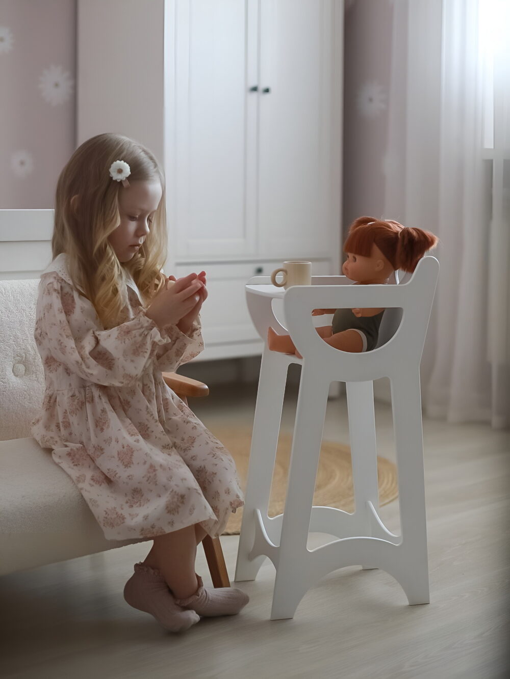 На стульчике для кормления кукол сидит игрушка, девочка в красивом розовом платье подает ей чат