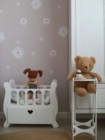 В комнате с обоями в цветочек, стоят белая люлечка для кукол и белый стульчик для кормления кукол с плюшевым медведем.