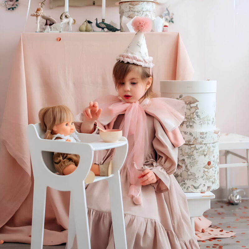Девочка в красивом розовом платье и карнавальном колпаке играет с куклой, которая сидит в стульчике для кормления кукол