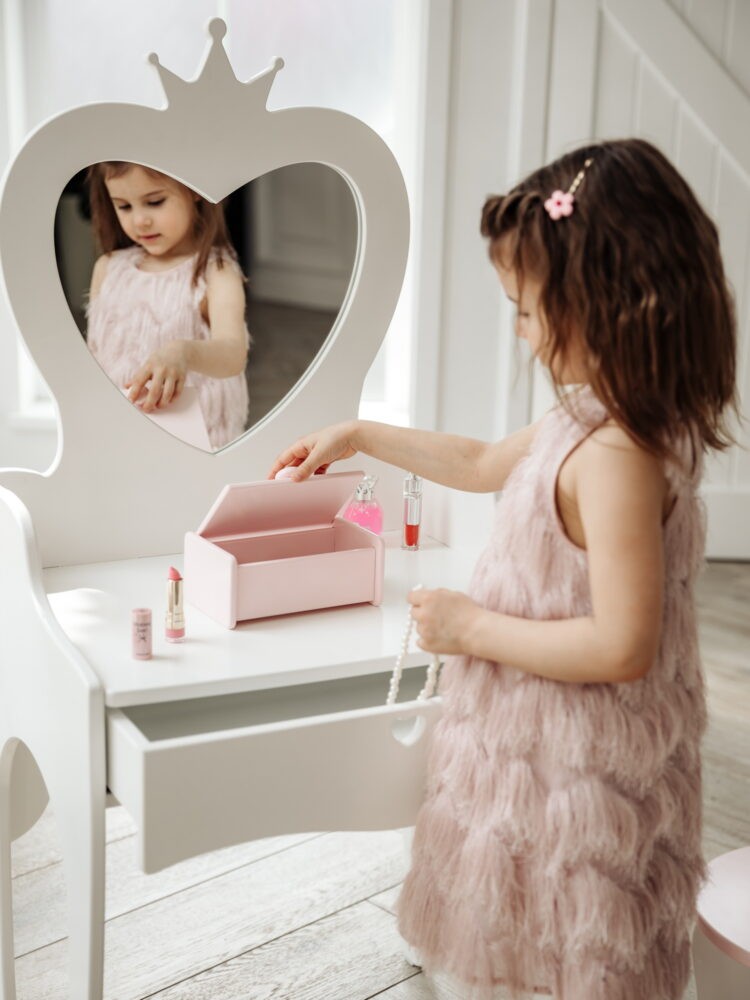 Девочка в красивом розовом платье стоит рядом с детским туалетным столиком с зеркалом и проверяет бижутерию в розовой детской шкатулке