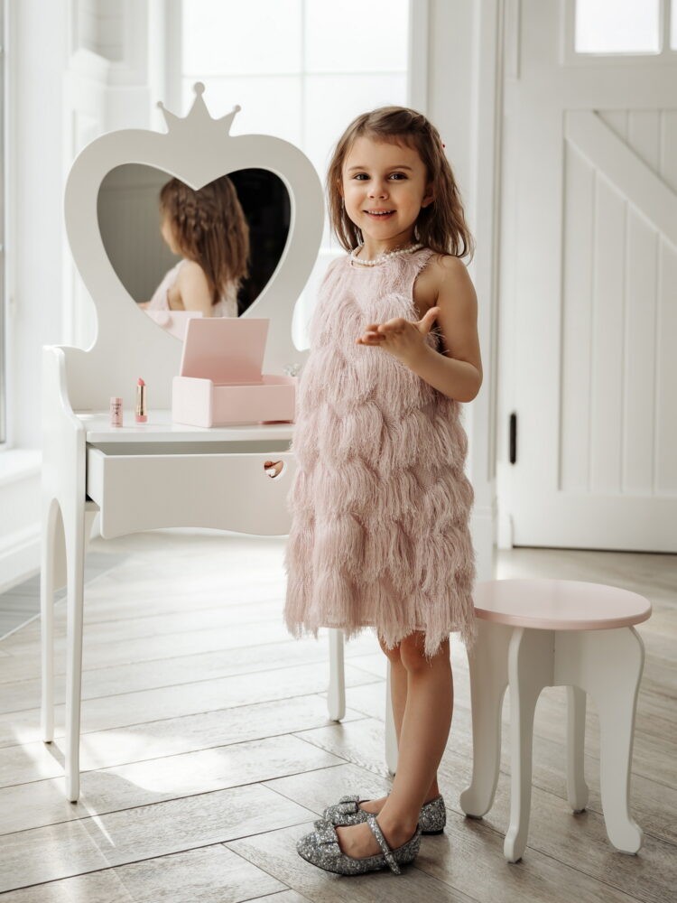 Девочка в красивом розовом платье стоит рядом с детским туалетным столиком с зеркалом и улыбается в камеру.