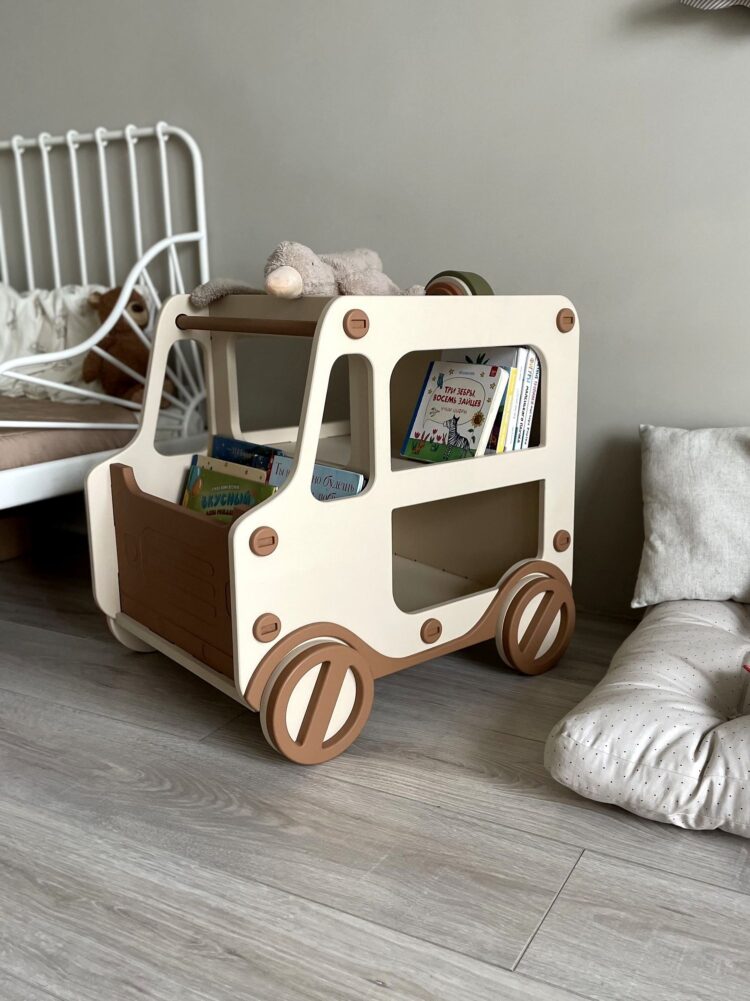 Рядом с кроватью в детской комнате стоит бежево-коричневая прикроватная тумба, полка для книг и игрушек в форме машины. Рядом лежит подушка, внутри тумбы книжки и игрушки