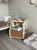 Рядом с кроватью в детской комнате стоит бежево-коричневая прикроватная тумба, полка для книг и игрушек в форме машины. Рядом лежит подушка, внутри тумбы книжки и игрушки