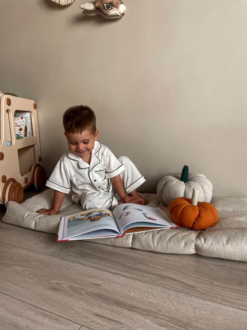 На пледе сидит мальчик и читает книжку рядом с бежево-коричневой прикроватной тумбой, полкой для книг и игрушек в форме машины